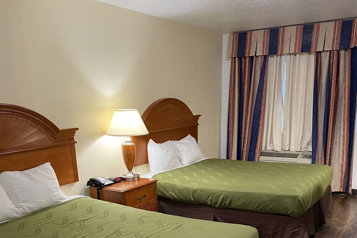 A hotel room with two beds, a nightstand with a lamp, a telephone, and a window with curtains.