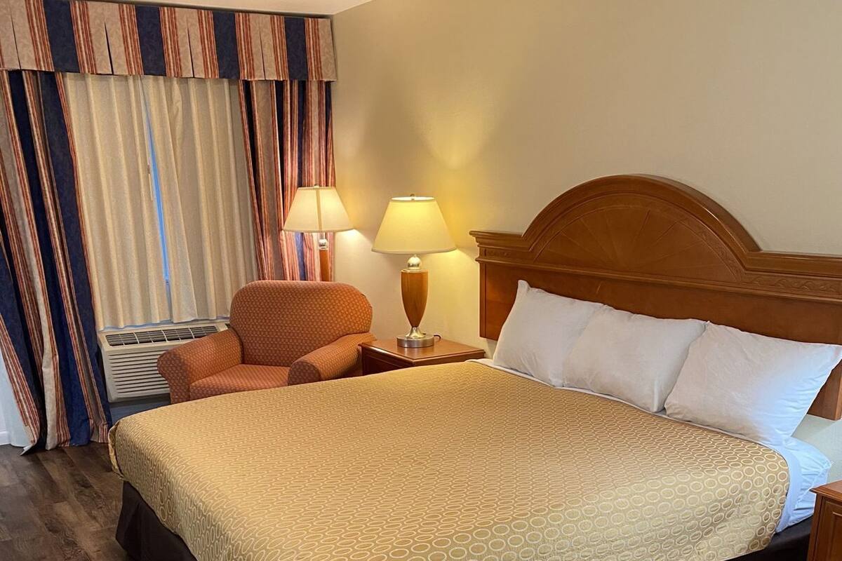 A hotel room with a large bed, a wooden headboard, a nightstand with a lamp, a chair, and a window with curtains.