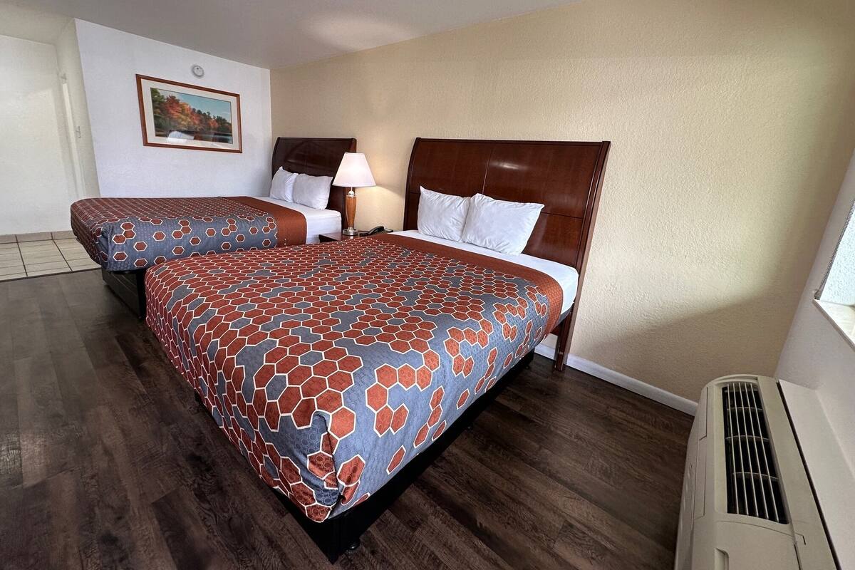 A hotel room with two beds, a nightstand, a lamp, a painting on the wall, and a window.