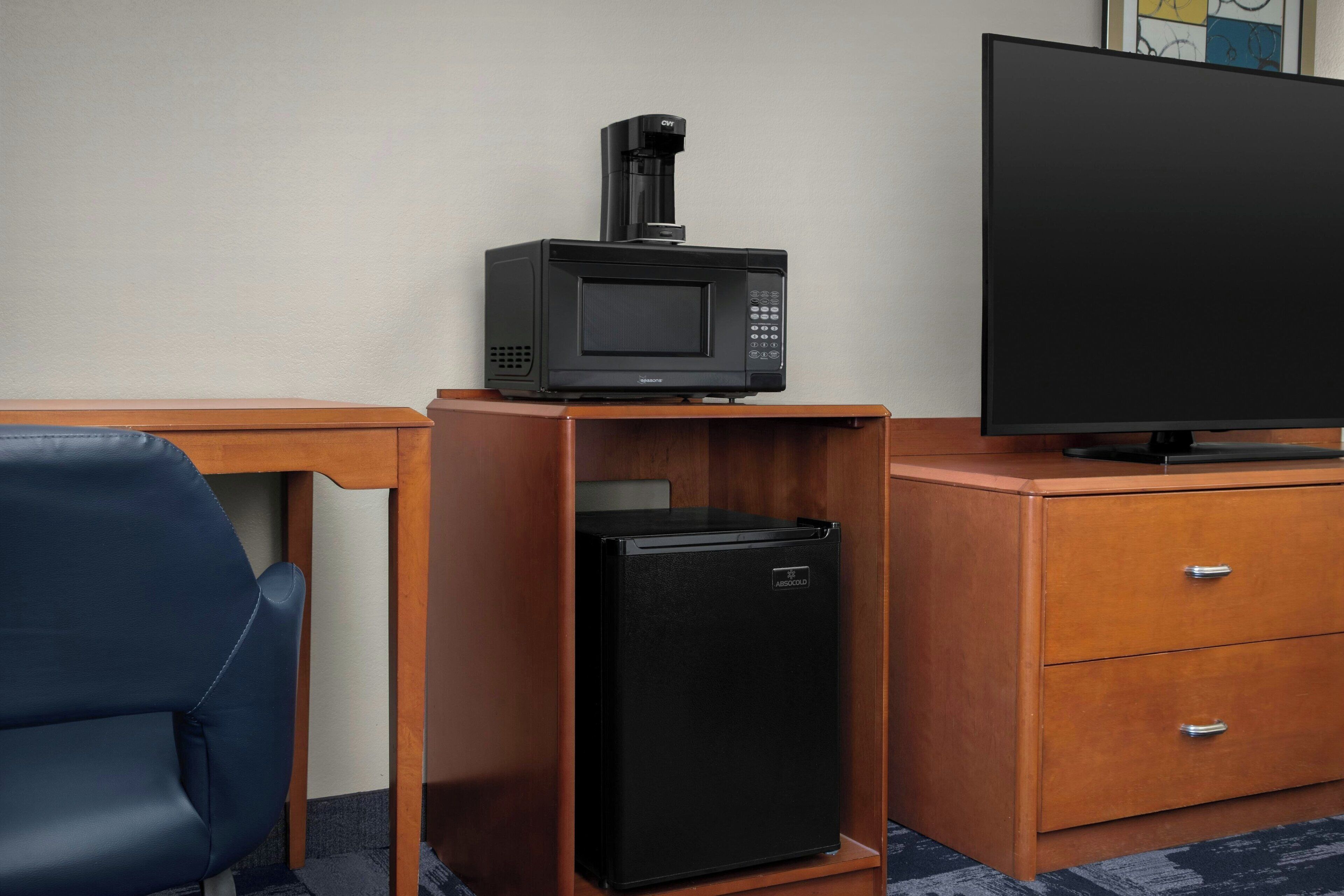 A compact kitchenette with a microwave, a small refrigerator, a television, and a wooden desk.