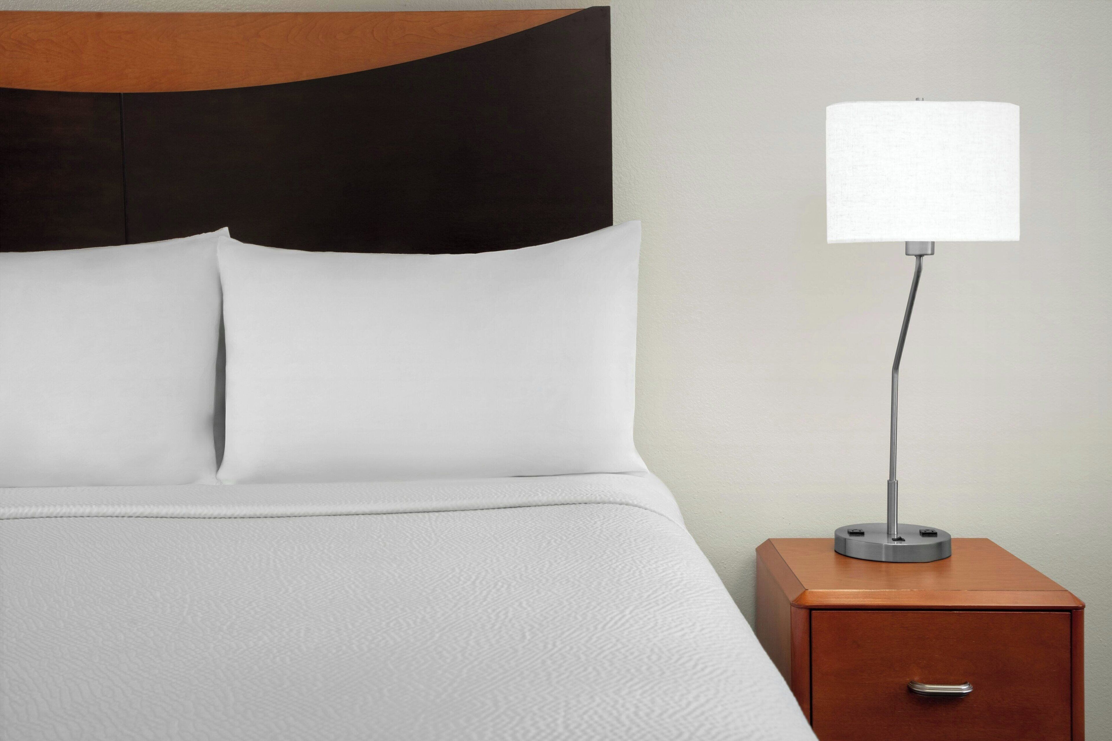 A neatly made bed with white linens and a wooden headboard, a bedside table with a lamp, and a drawer.