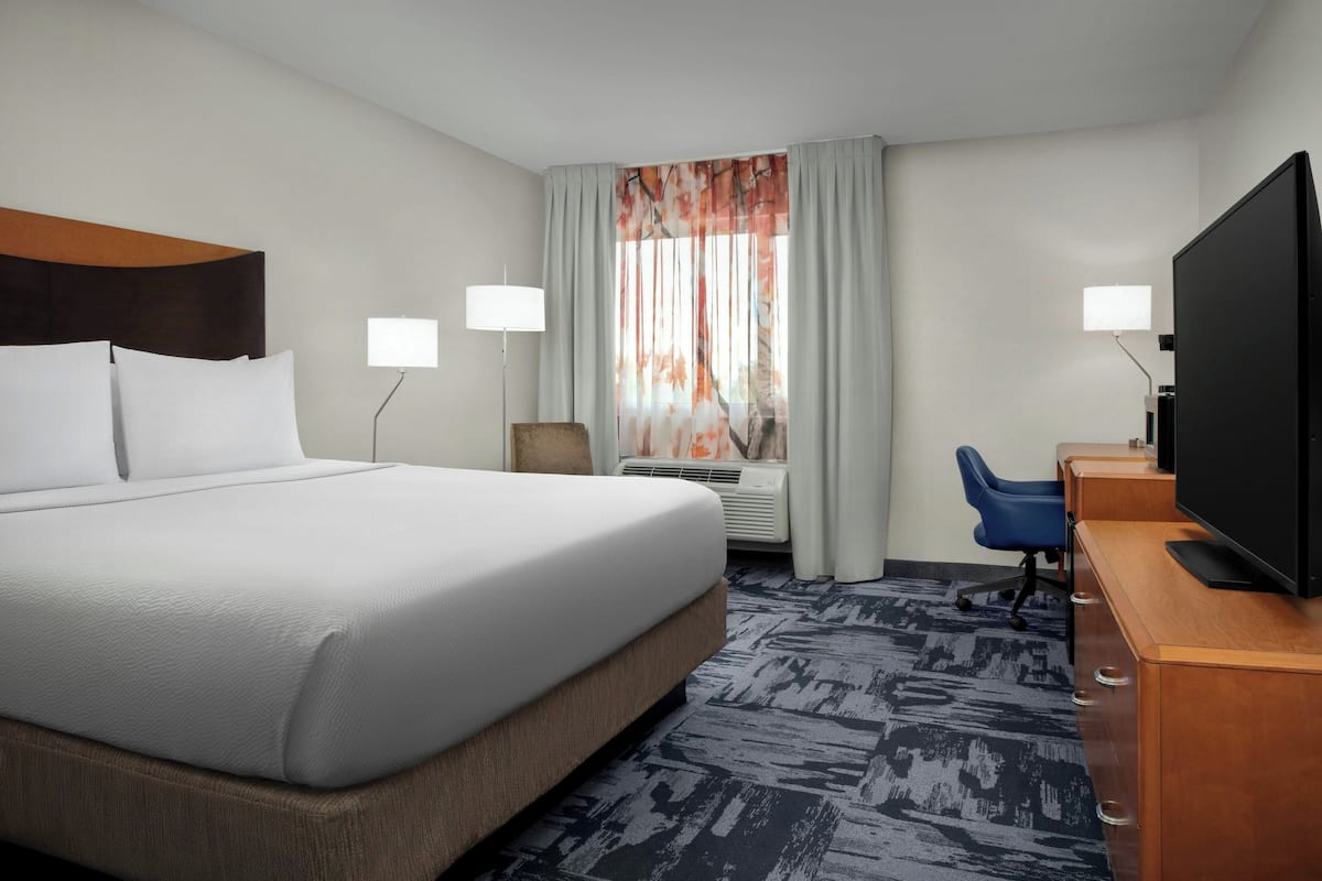A hotel room with a large bed, a desk with a flat-screen TV, a chair, and a window with curtains.