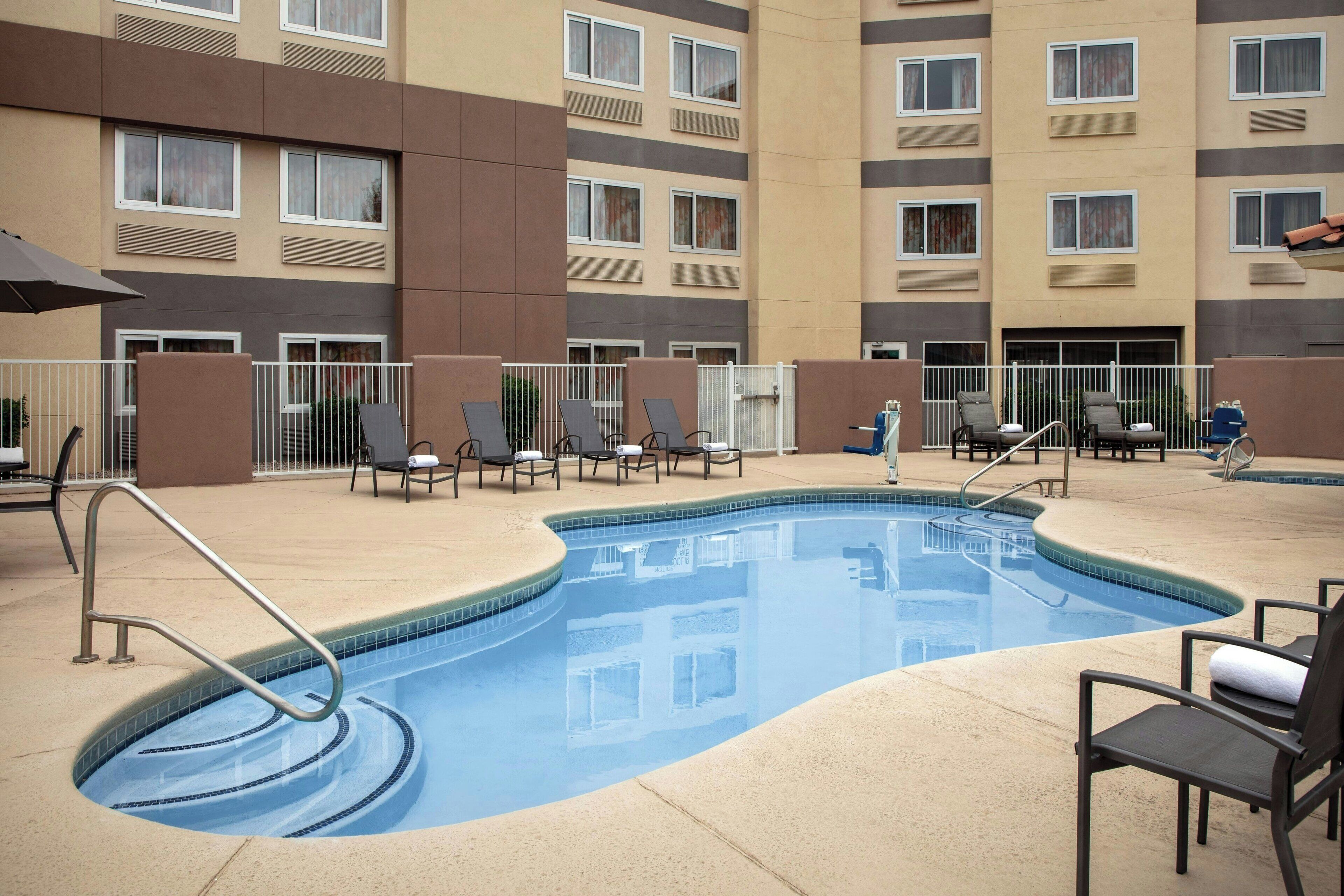 A swimming pool with lounge chairs and a fence, surrounded by a multi-story building with balconies.