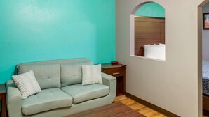 A room with teal walls, a grey sofa with patterned cushions, a wooden nightstand, and a mirror above a wardrobe. - Econo Lodge West - Coors Blvd (Albuquerque)