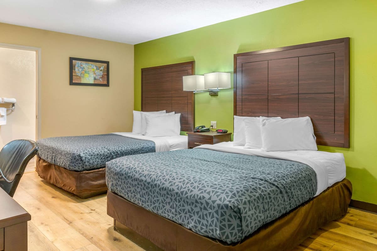 Habitación de hotel con dos camas, un escritorio, una silla, una mesita de noche y una lámpara de pared.