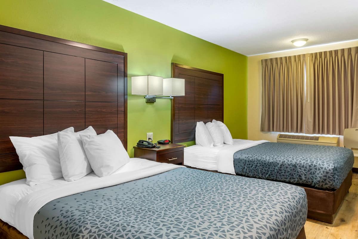 Habitación de hotel con dos camas, una mesita de noche con lámpara, una ventana con cortinas y un aire acondicionado de pared.