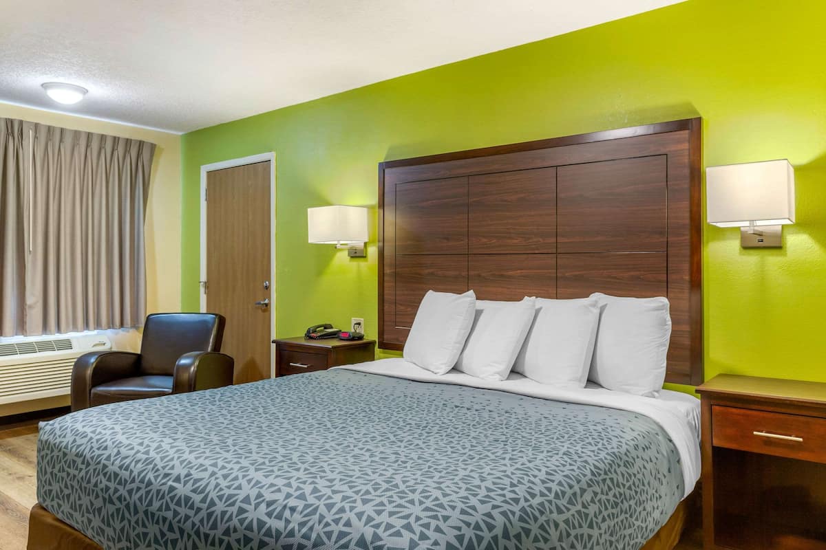 Una habitación de hotel con una cama grande, un cabecero de madera, una silla, una mesita de noche, una lámpara y una ventana con cortinas.