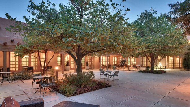 An outdoor patio area with seating arrangements, trees, and a building with large windows. - Courtyard by Marriott Albuquerque (Albuquerque)