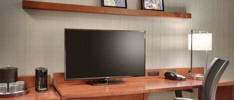 A hotel room with a wooden desk, a flat-screen TV, a coffee maker, a lamp, and a chair.