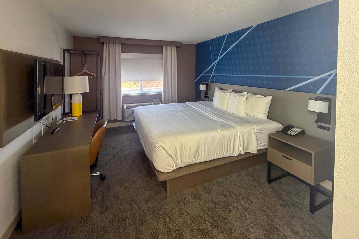 A hotel room with a bed, a desk, a TV, a chair, a window with curtains, and a wall with a geometric pattern.