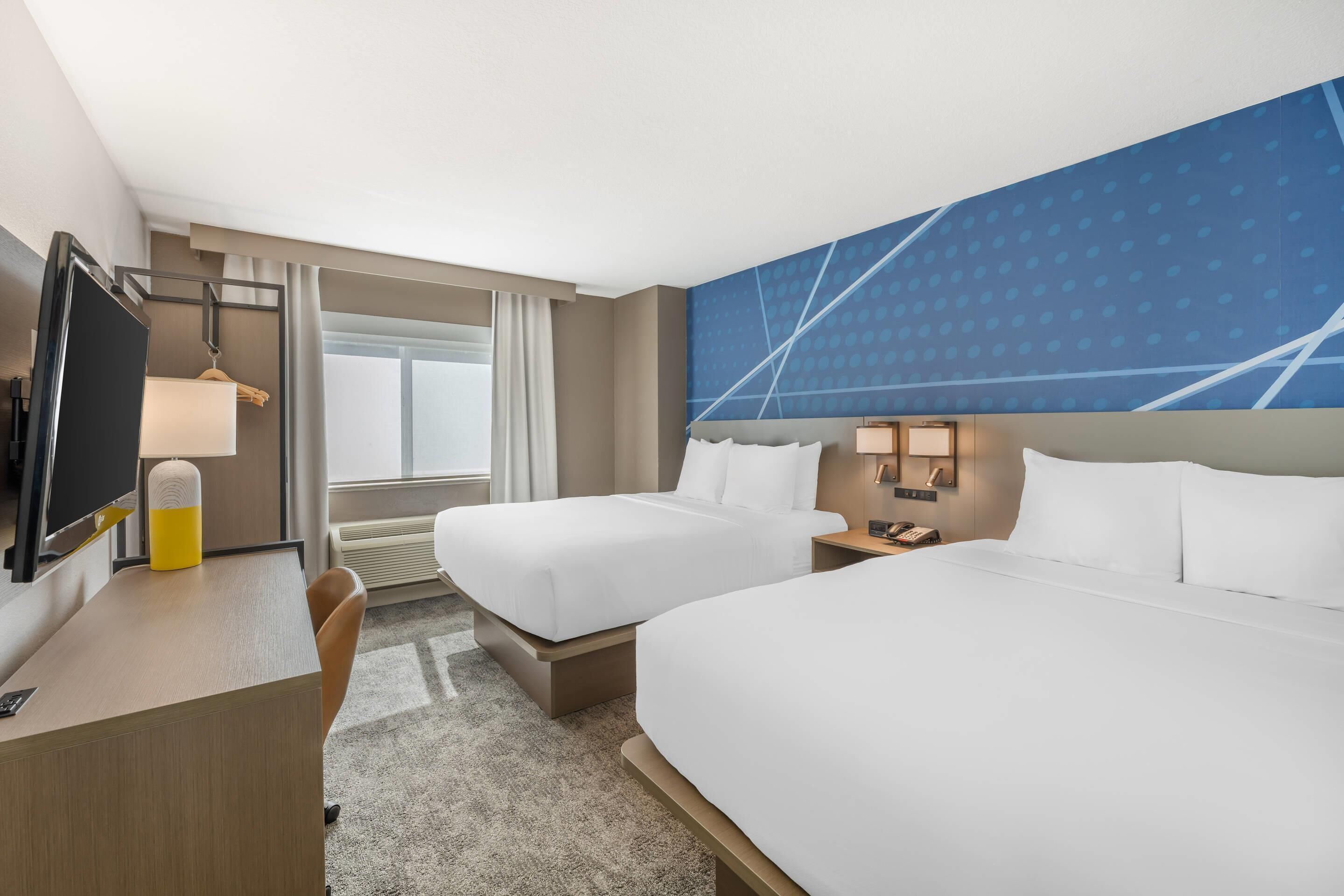 A hotel room with two beds, a desk with a chair, a television, and a large window with curtains.
