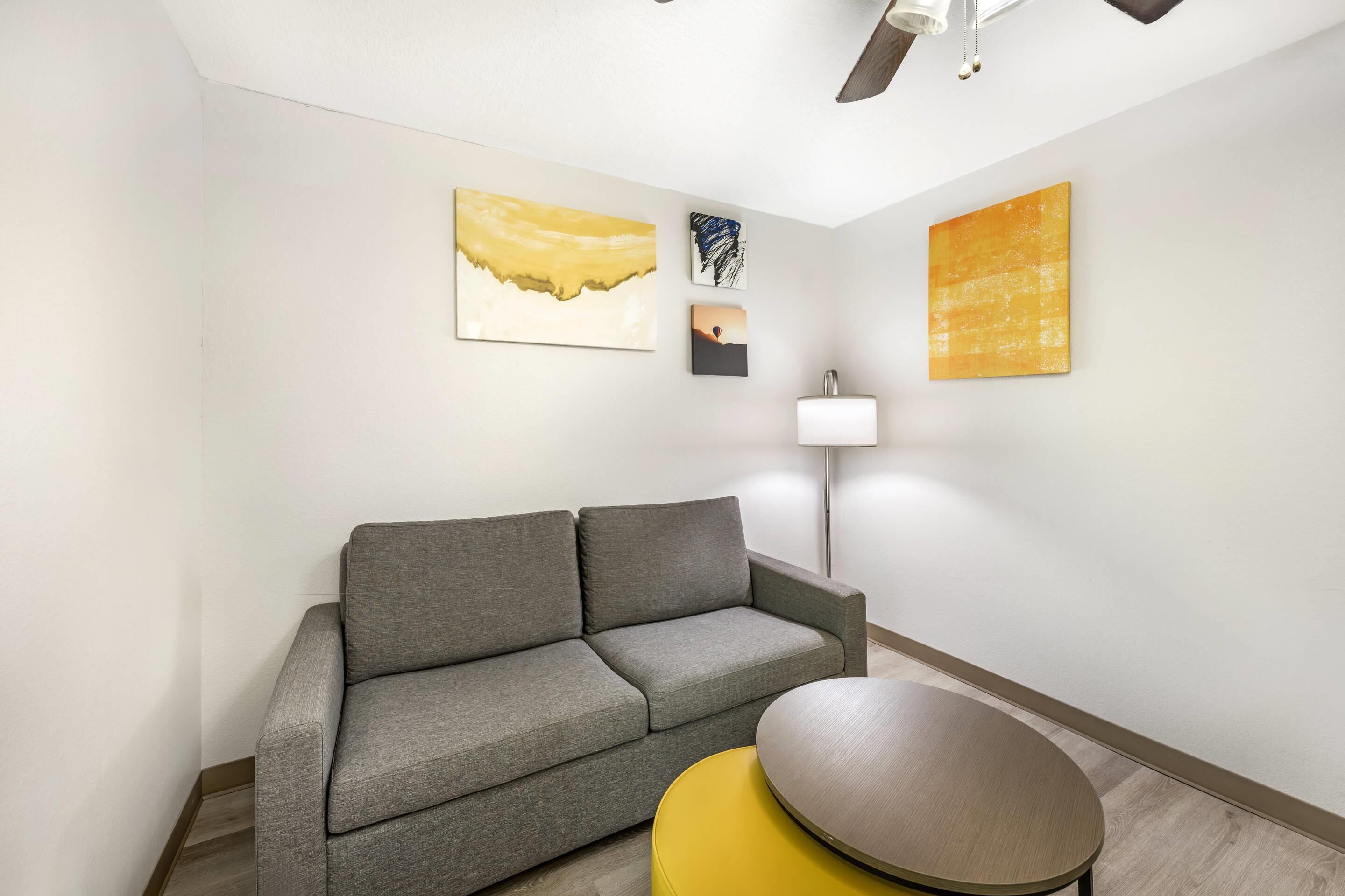 Ein modernes Wohnzimmer mit einem grauen Sofa, einem gelben Pouf und einem runden Tisch. An der Wand hängen drei Kunstwerke.