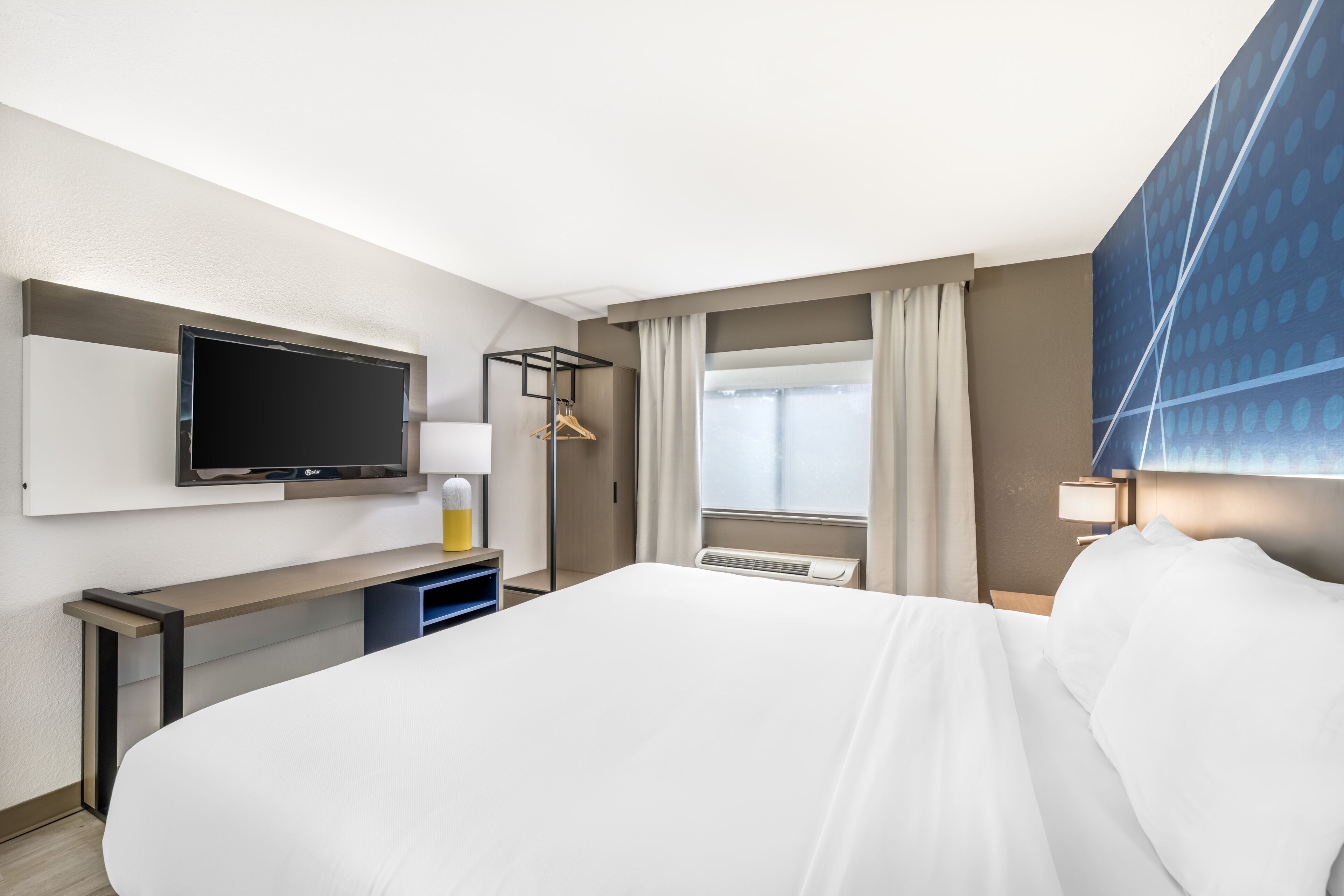 Une chambre d’hôtel avec un lit, un téléviseur fixé au mur, un bureau avec une lampe et une fenêtre avec des rideaux.