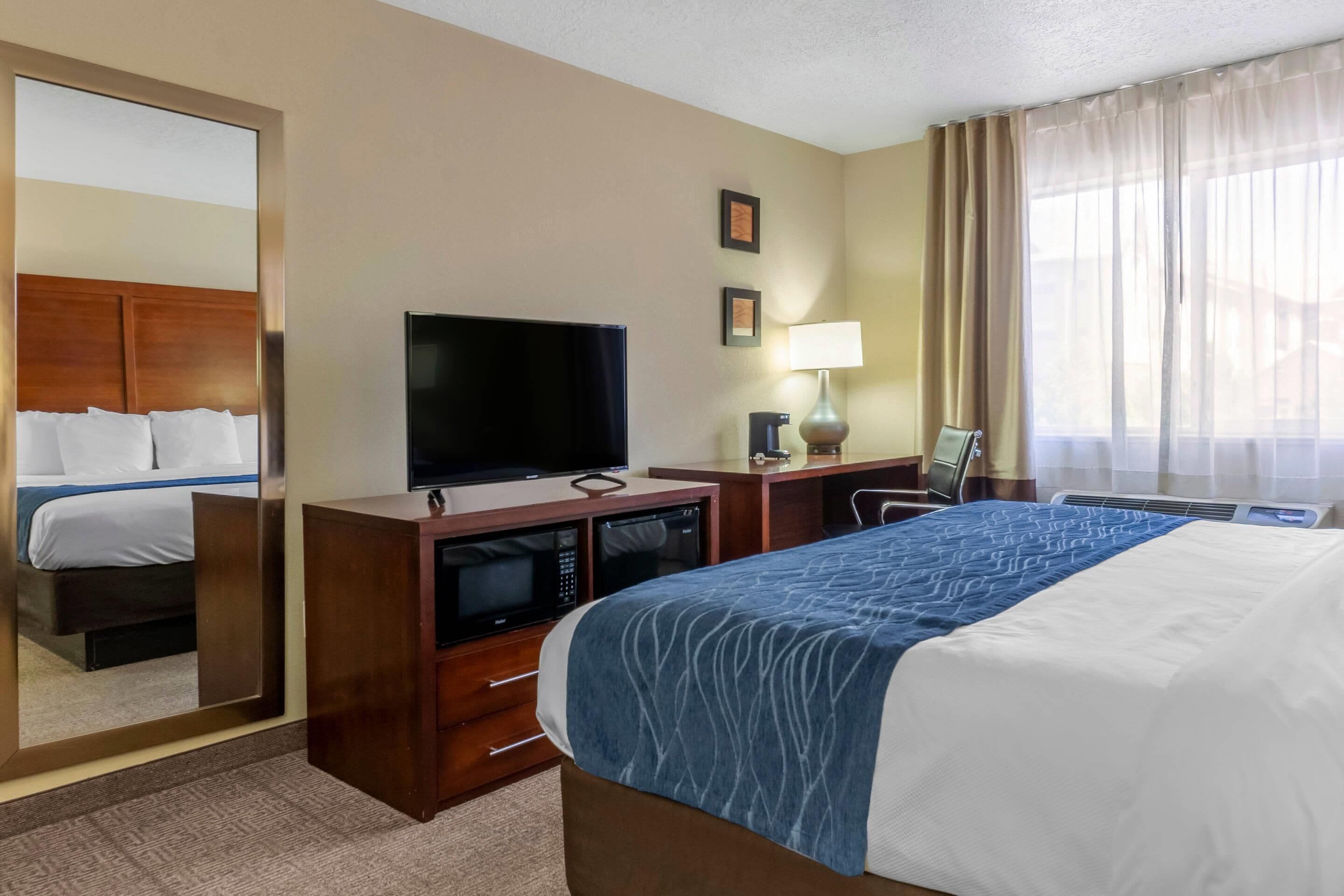 A hotel room with a bed, a TV, a desk with a lamp, and a large mirror.