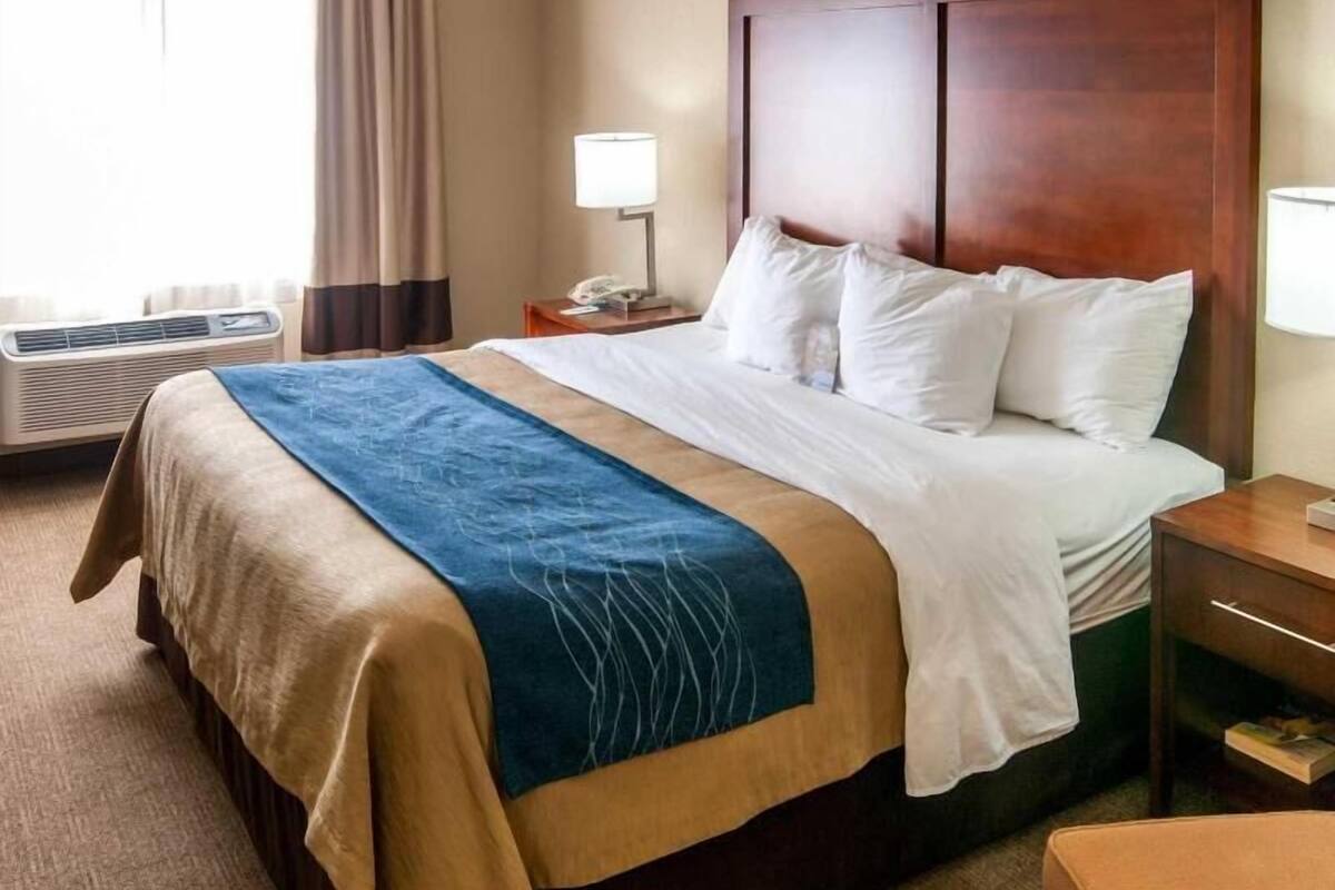 Ett hotellrum med en säng, ett nattduksbord, en lampa, en luftkonditioneringsanläggning och ett fönster med gardiner.