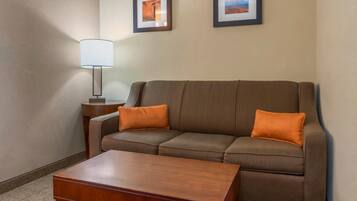 Une chambre d’hôtel avec un canapé marron, deux coussins orange, une table basse en bois, une lampe et deux tableaux encadrés accrochés au mur.