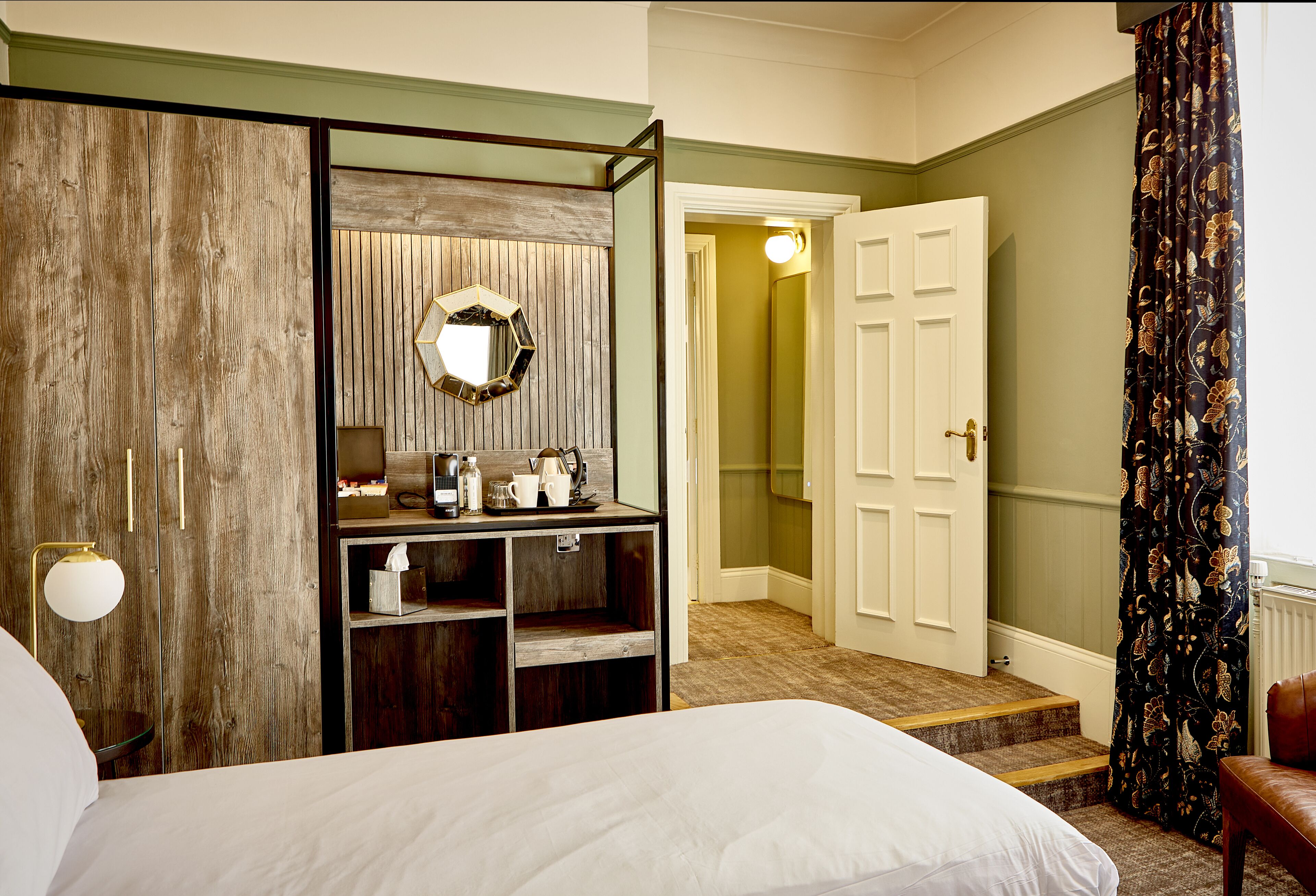Une chambre d’hôtel comprenant un lit, une armoire en bois, un miroir, une étagère avec des articles de toilette et une porte donnant sur une autre pièce.