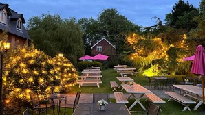 Un patio al aire libre con mesas y sillas de picnic, rodeado de árboles y una casa al fondo.