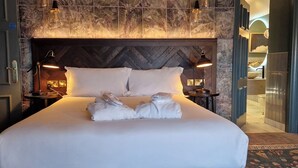 Una cama bien hecha con sábanas y toallas blancas, un cabecero de madera, dos lámparas de noche y vistas a un baño con lavabo e inodoro.