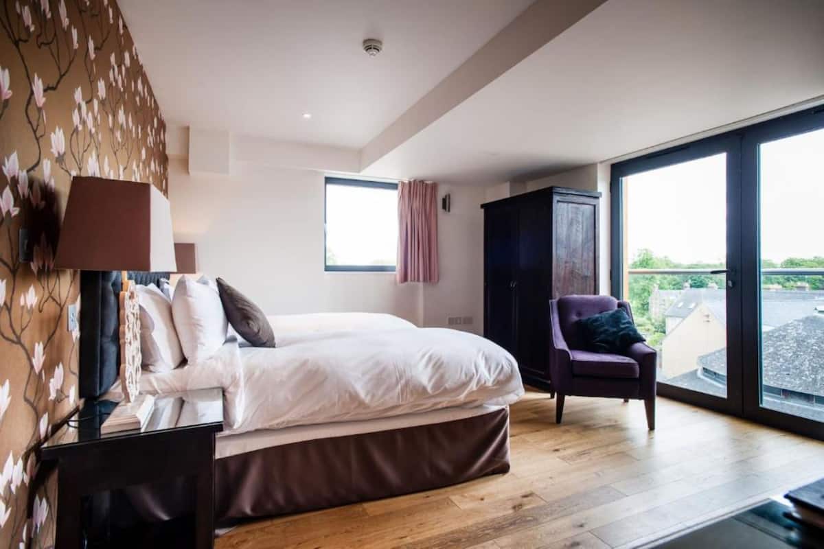 A bedroom with a bed, a chair, a wardrobe, a window, and a view of the outside.