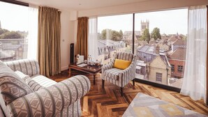 A spacious living room with a large window offering a city view, a patterned sofa set, a small armchair with a yellow cushion, and a herringbone-patterned rug. - The Varsity Hotel & Spa (Cambridge)