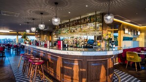 A bar area with a wooden bar counter, multiple stools, hanging crystal chandeliers, and a view of the cityscape through large windows. - The Varsity Hotel & Spa (Cambridge)
