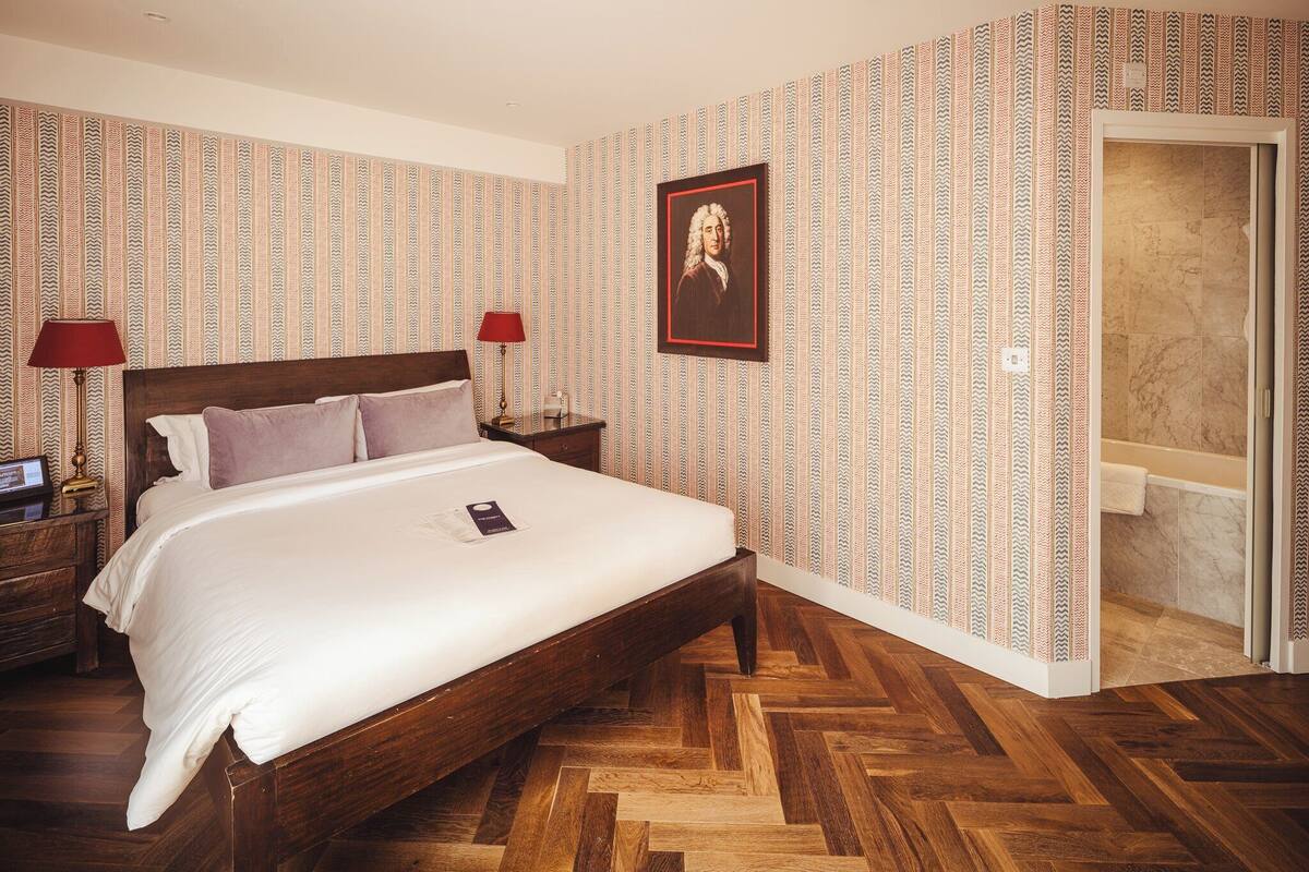 A hotel room with a wooden herringbone floor, a large bed with white linens, two bedside tables with lamps, and a patterned wallpaper.