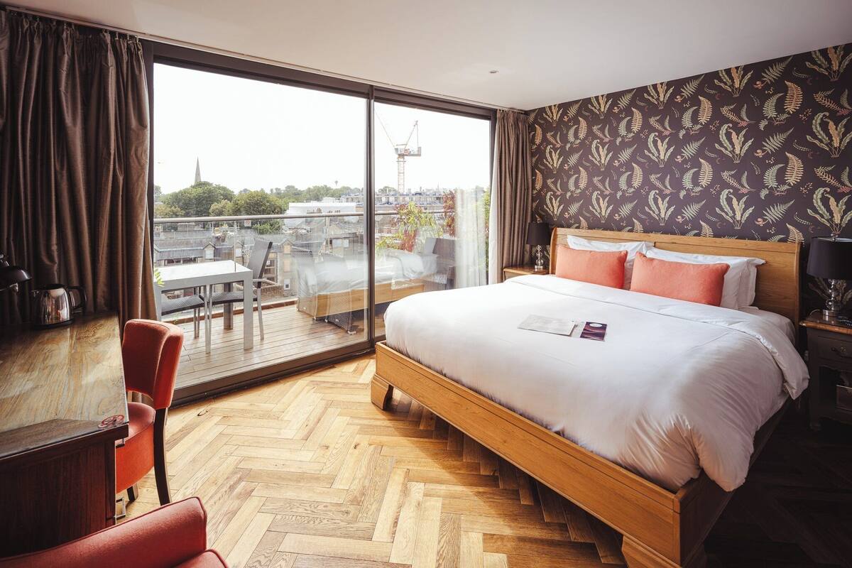 A hotel room with a large bed, a wooden floor, a balcony with a table and chairs, and a view of the cityscape.