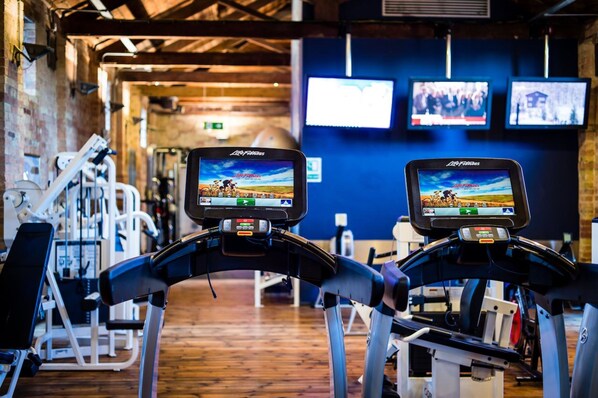 A gym with treadmills displaying a movie, a brick wall, and exercise equipment.