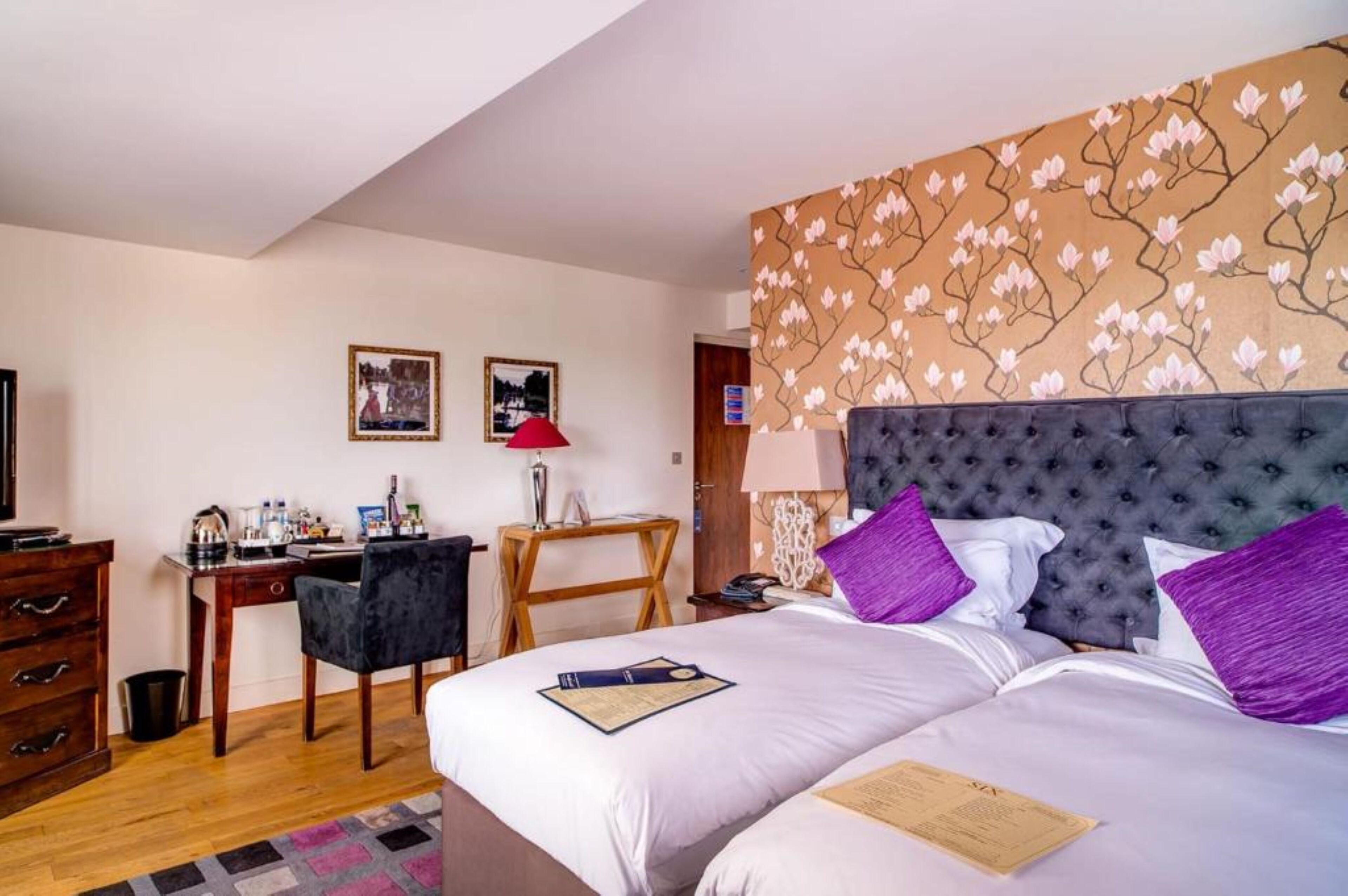 A hotel room with a large bed, a desk, a chair, a dresser, and a wall with a floral pattern.