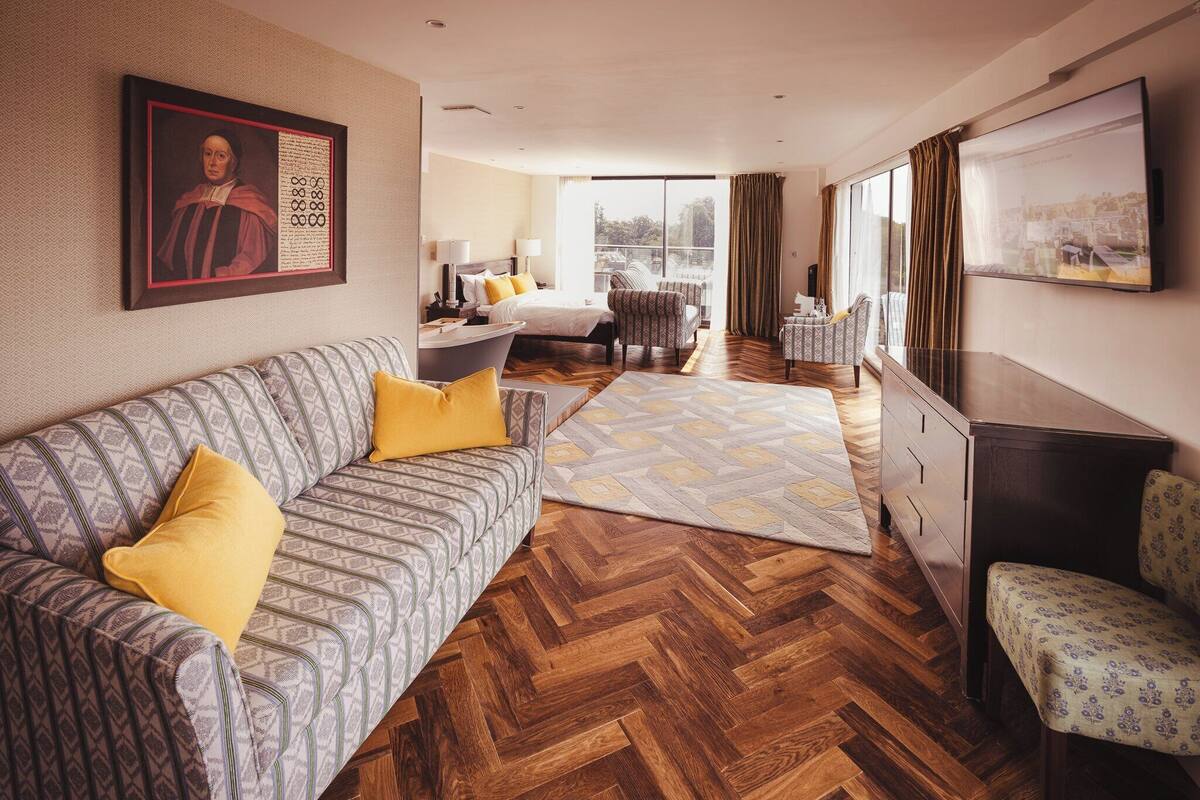 A hotel room with a patterned sofa, a herringbone wooden floor, a large window with curtains, a flat-screen TV, and a painting on the wall.