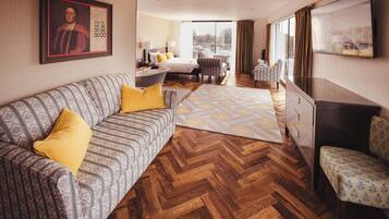 A hotel room with a patterned sofa, a herringbone wooden floor, a large window with curtains, a flat-screen TV, and a painting on the wall.
