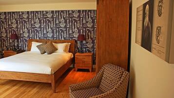 A bedroom with a wooden bed, patterned wallpaper, a nightstand, a chair, and a wall-mounted artwork.