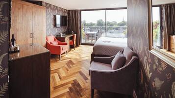 Habitación de hotel con piso de madera tipo tablero de ajedrez, una cama, un escritorio con silla, un sillón rojo y un televisor.