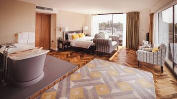 A modern hotel room with a large bathtub, a bed with yellow pillows, a seating area with patterned chairs, and a large window offering a view.