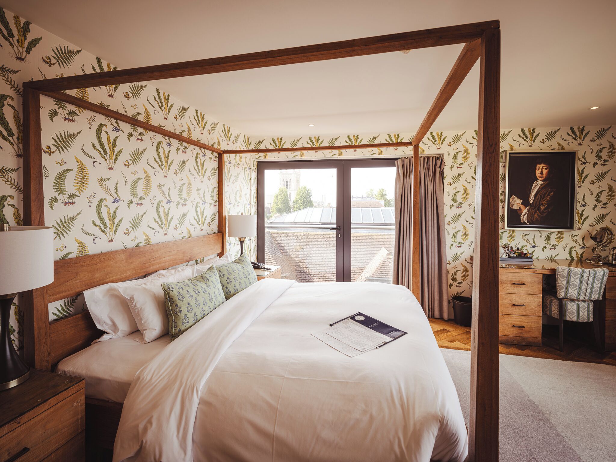 A four-poster bed with a wooden headboard, a nightstand with a lamp, a desk with a chair, and a large window with curtains.