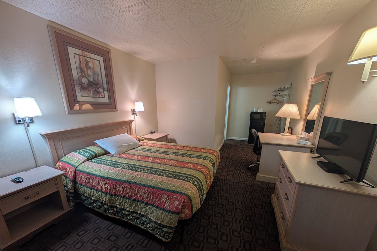 A hotel room with a bed, bedside table, lamp, desk with a chair, TV, and a mirror.