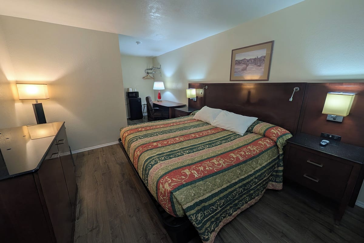 A hotel room with a bed, bedside tables, a desk, a chair, a desk lamp, a wall-mounted TV, and a painting on the wall.