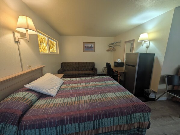 A hotel room with a bed, a sofa, a desk with a computer, a refrigerator, and wall-mounted lamps.