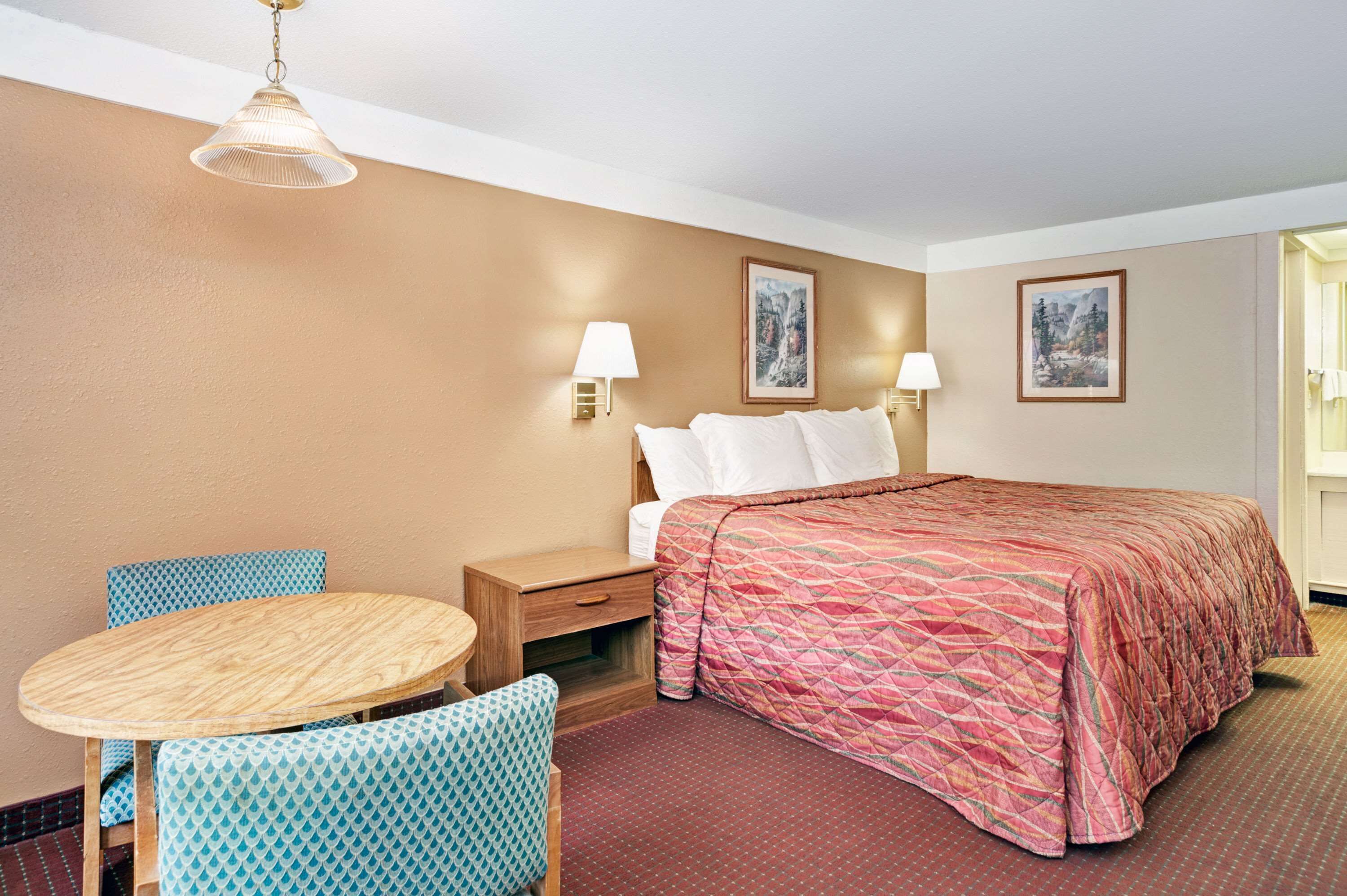 A hotel room with a bed, two bedside lamps, a round wooden table, two patterned chairs, and two wall-mounted paintings.