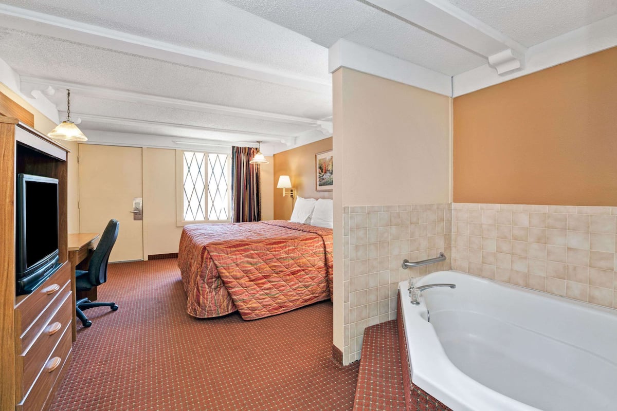 A hotel room with a bed, a desk with a television, a bathroom with a bathtub, and a window with curtains.