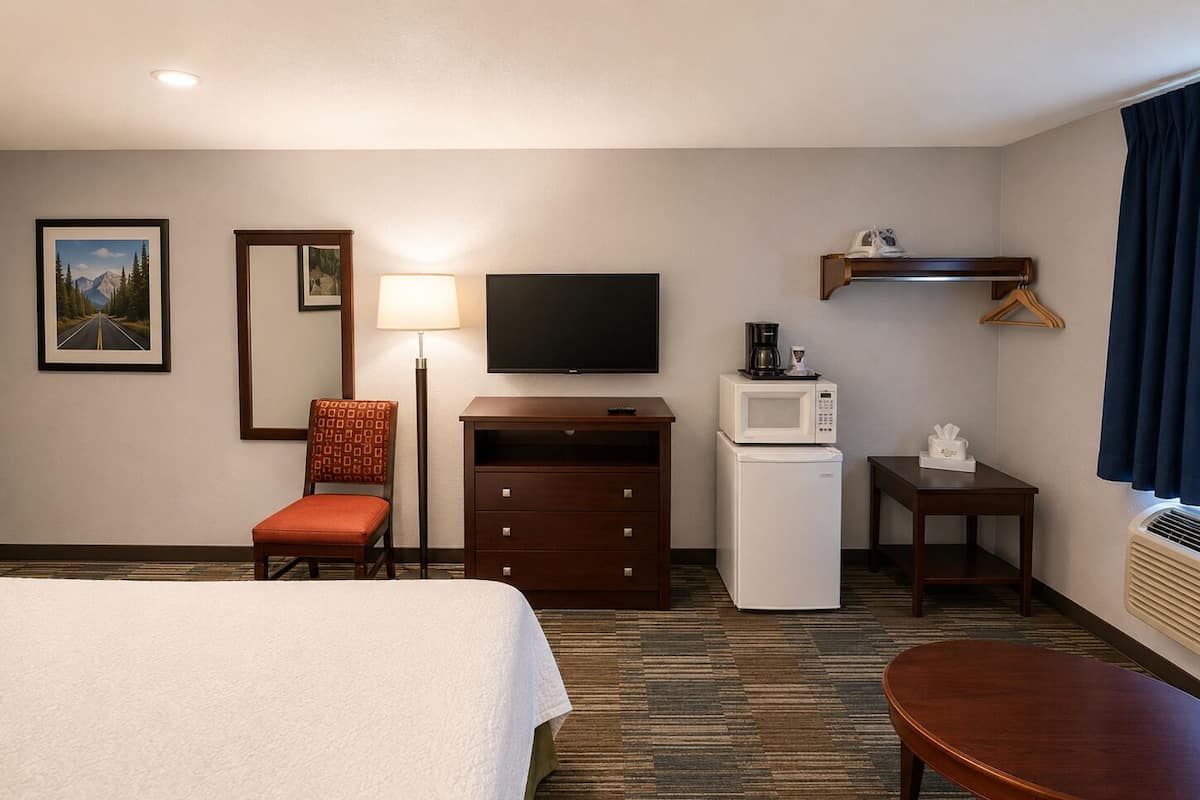 A hotel room with a bed, a desk, a chair, a television, a microwave, a small table, and a picture on the wall.