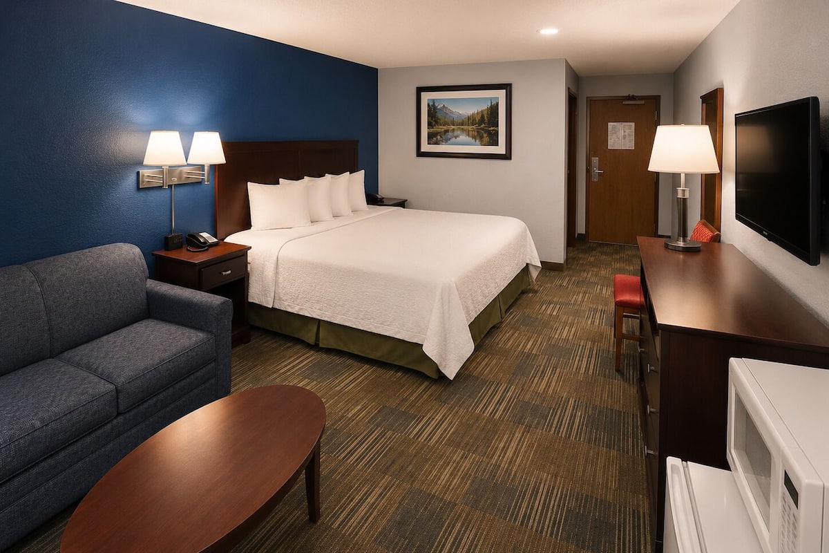 A hotel room with a bed, a sofa, a desk, a television, and a painting on the wall.
