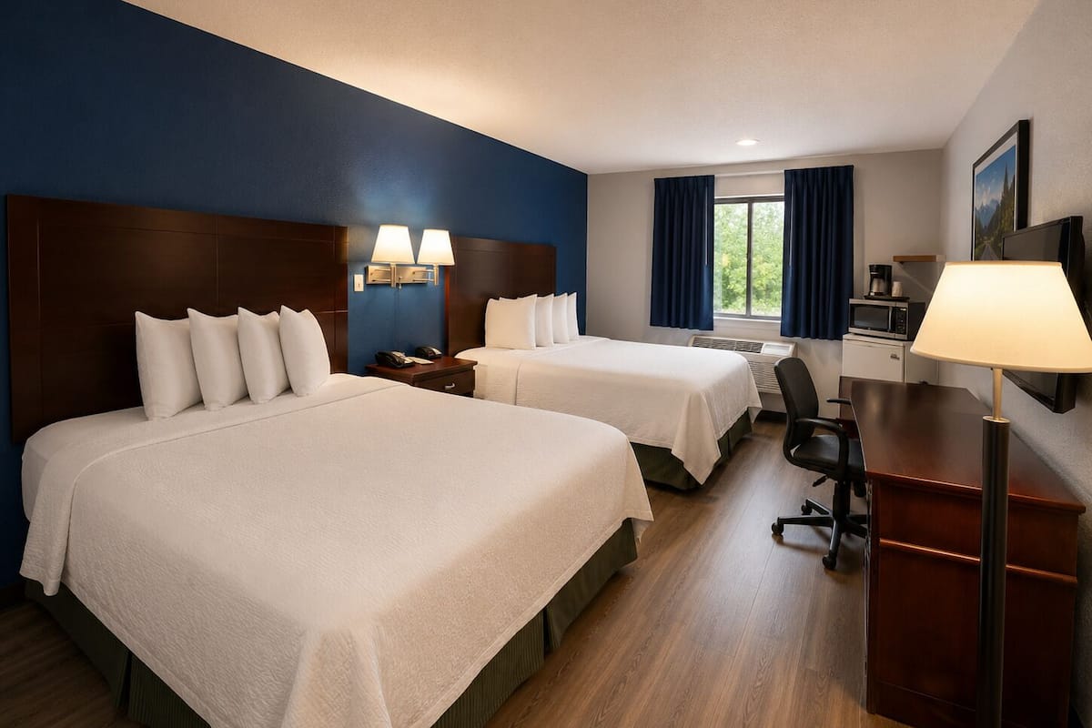 A hotel room with two beds, a desk, a chair, a microwave, and a TV.