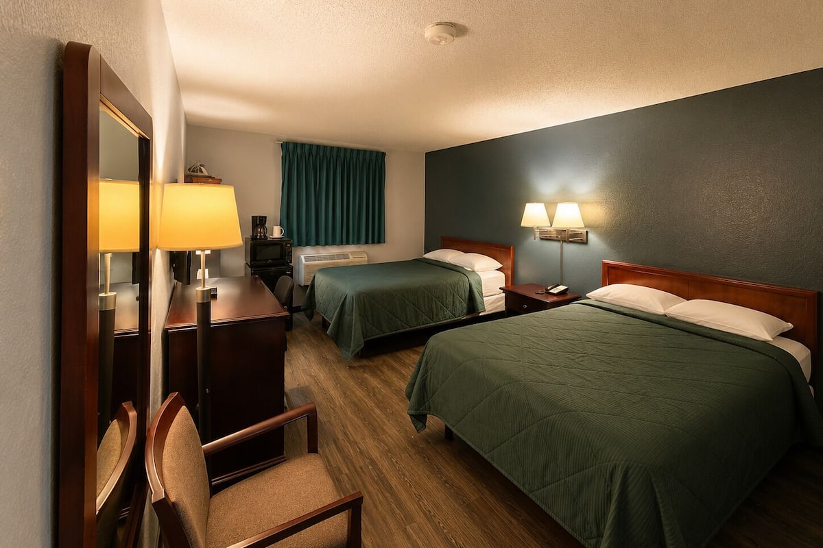 A hotel room with two beds, a desk with a chair, a mirror, a television, and a window with curtains.