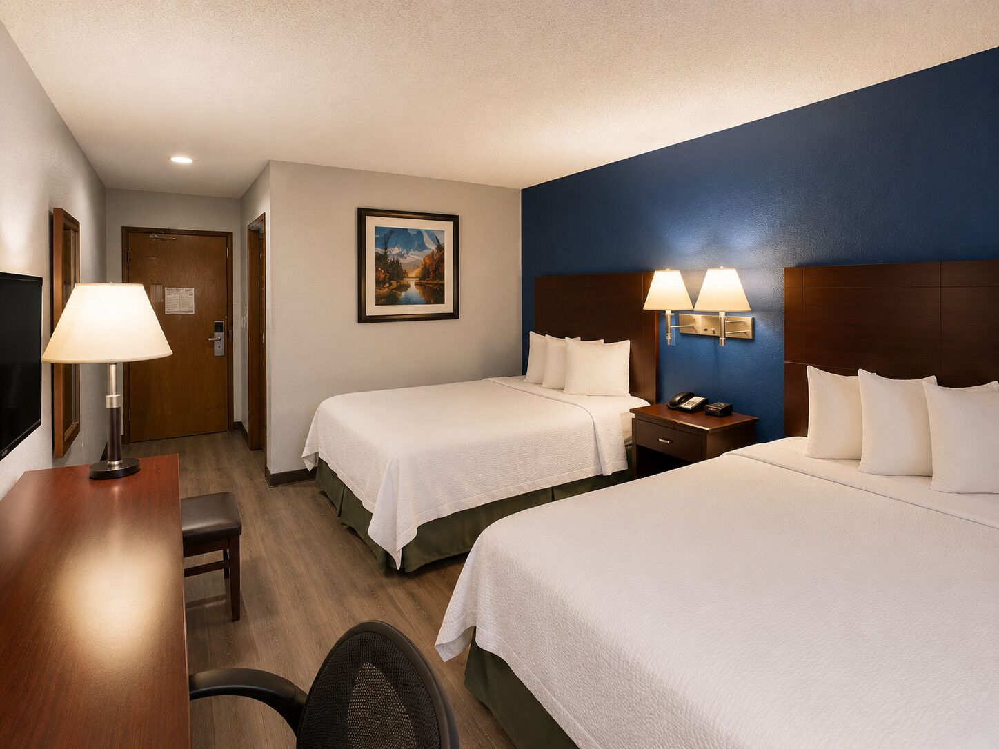 A hotel room with two beds, a desk, a chair, a nightstand with a lamp, and a painting on the wall.