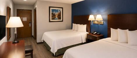 A hotel room with two beds, a desk, a chair, a nightstand with a lamp, and a painting on the wall.