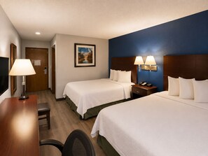 A hotel room with two beds, a desk, a chair, a nightstand with a lamp, and a painting on the wall.