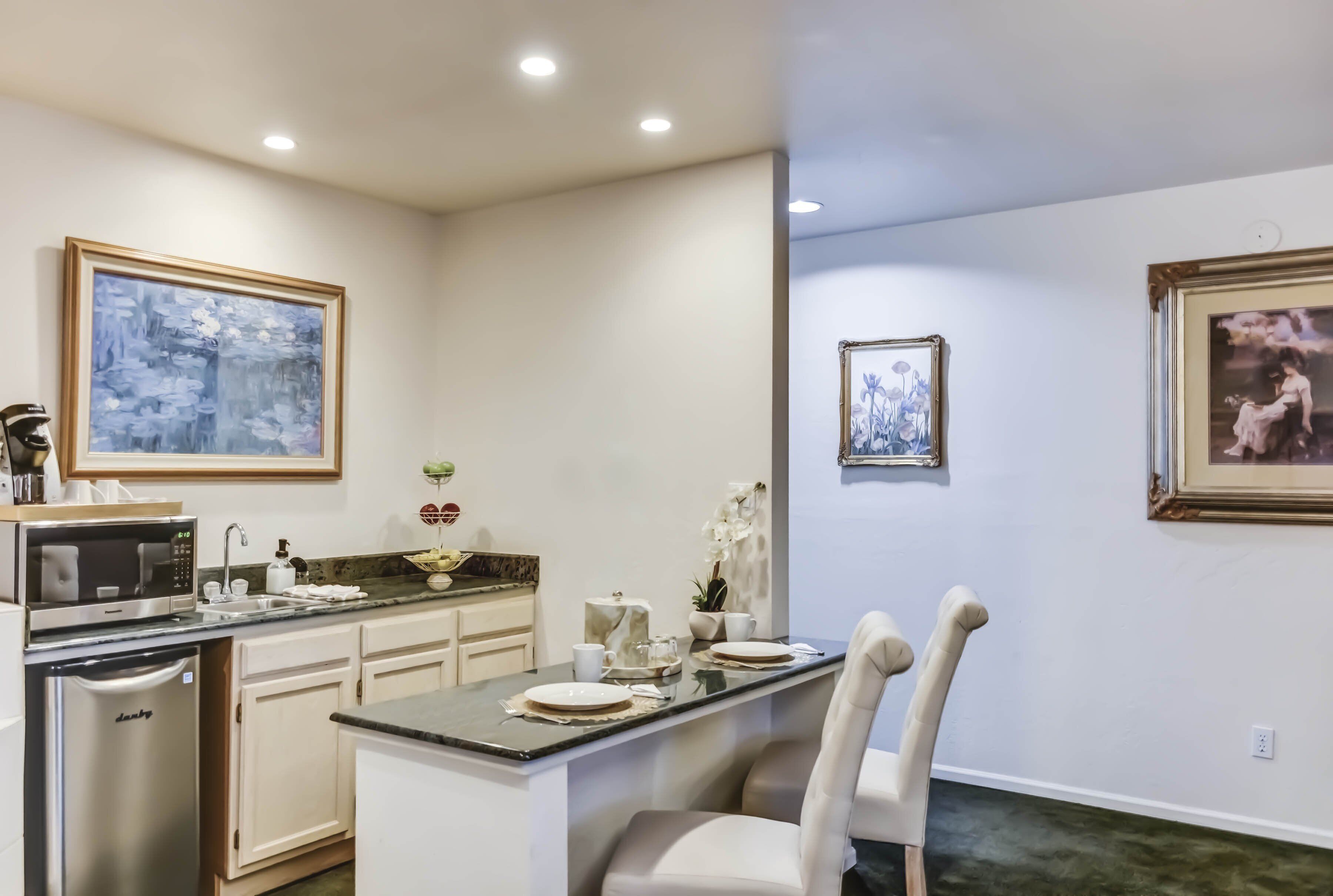 A modern kitchen with a central island, white cabinetry, and a microwave. There are framed artworks on the walls and a dining area with a chair.