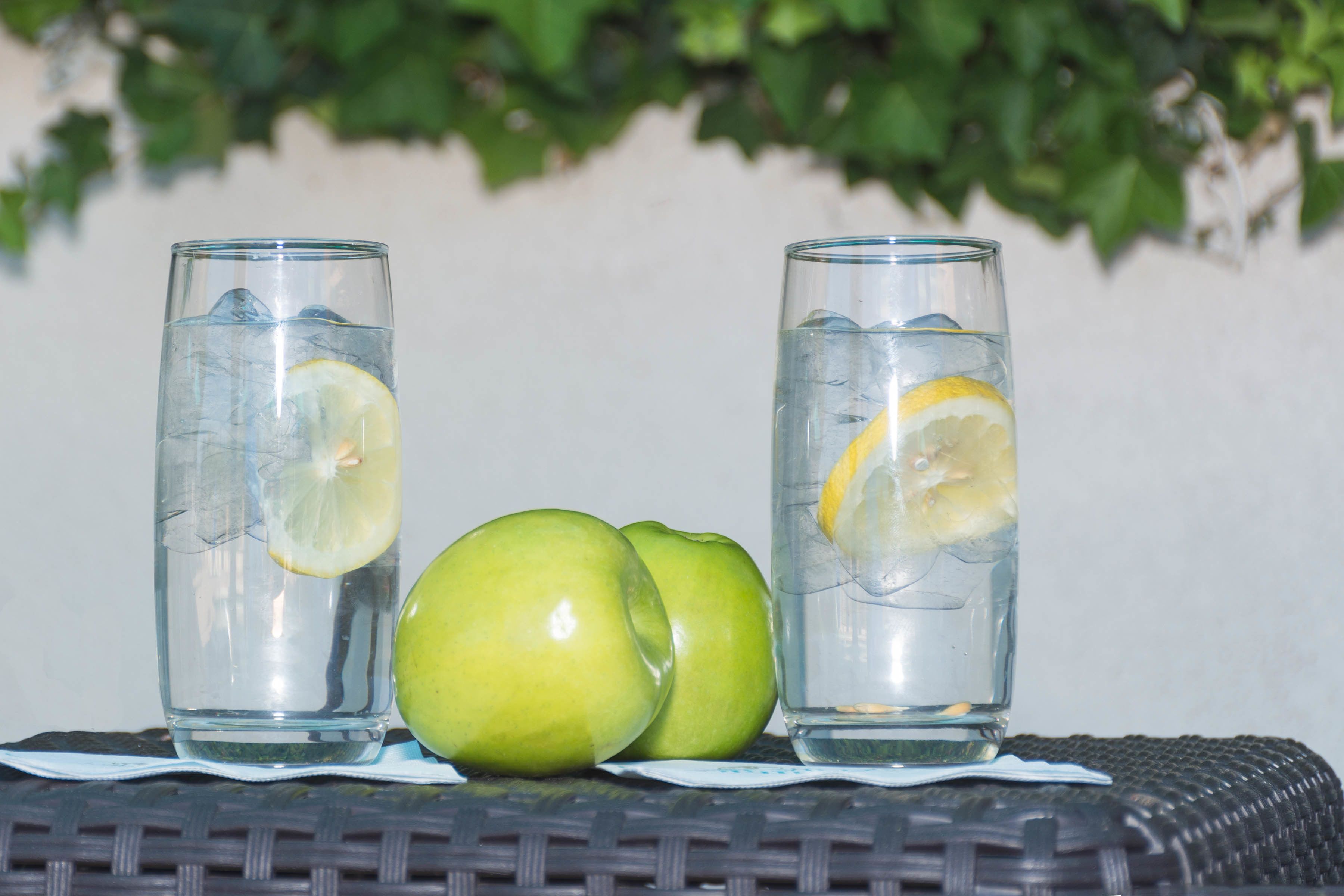 Dos vasos de agua con rodajas de limón y dos manzanas verdes sobre una mesa de mimbre.